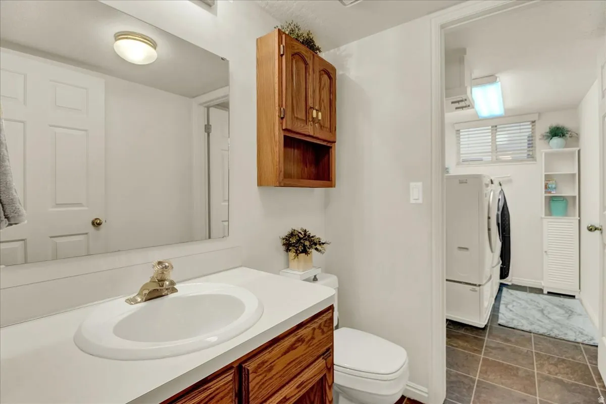 Bathroom featuring a wood-finish vanity with white countertop and integrated sink, large framed mirror, wall-mounted cabinetry with open shelving, ceramic tile flooring, and direct access to laundry area