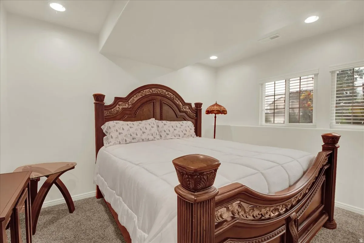 Elegant primary bedroom showcasing ornate wood-carved bed frame with upholstered headboard, recessed ceiling lighting, plantation shutters, and neutral wall finishes