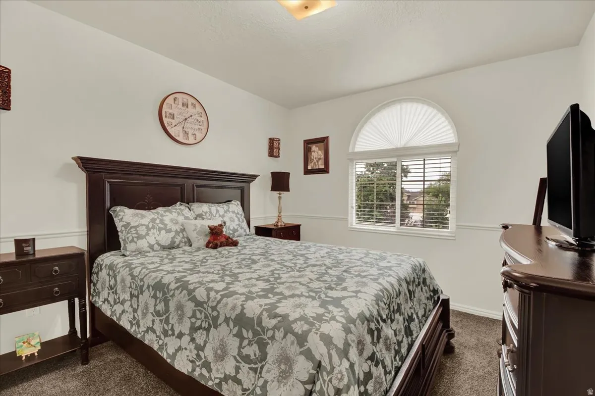 Spacious primary bedroom featuring dark wood furnishings, a decorative arched window with plantation shutters, neutral patterned bedding, carpeted flooring, and recessed ceiling lighting throughout the room