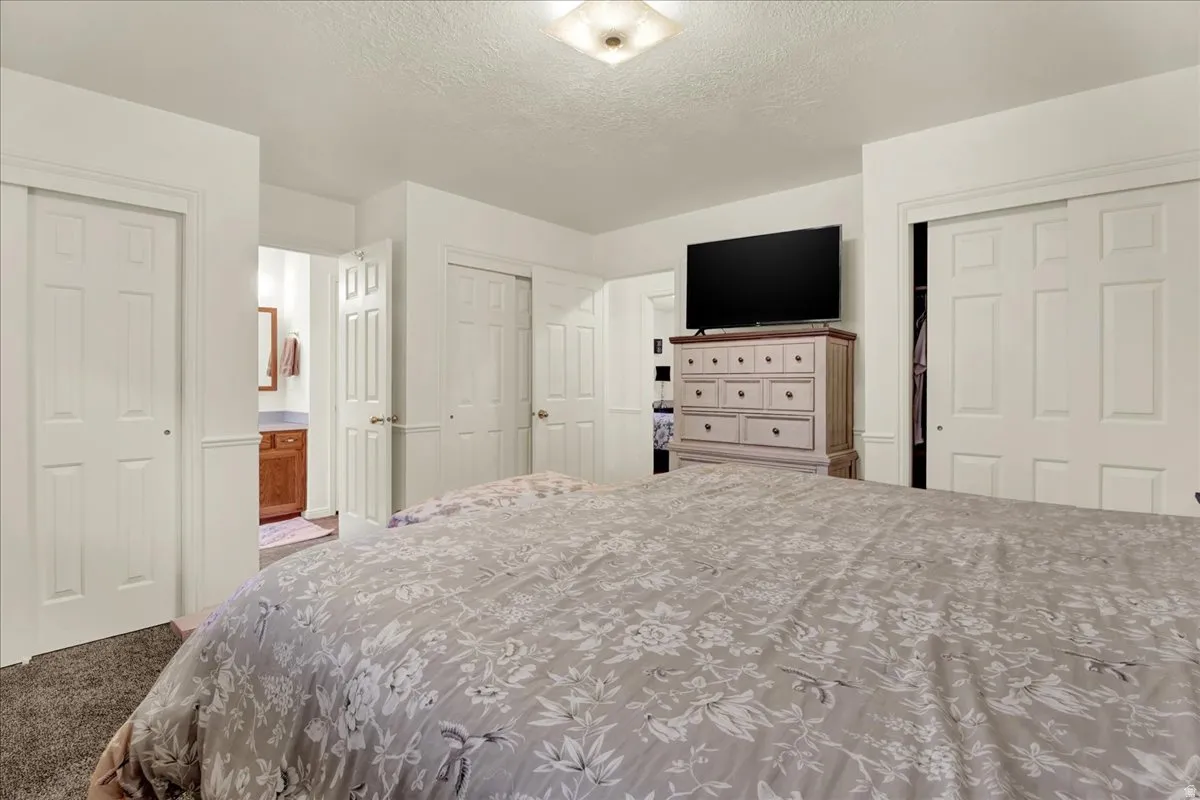 Spacious primary bedroom suite with multiple closet doors, carpeted flooring, wall-mounted flat-screen television, and wooden dresser with drawer storage
