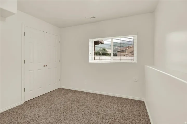 Bright secondary bedroom showcasing neutral wall finishes, carpet flooring, a double-door closet with white six-panel doors, and a horizontal window with exterior shutters allowing natural light