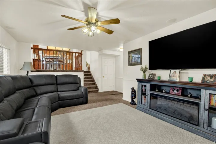 Spacious recreation room featuring a curved sectional sofa arrangement, wall-mounted flat-screen display, built-in media console with shelving and fireplace insert, ceiling fan with integrated lighting, neutral carpeting, and access to a wooden staircase