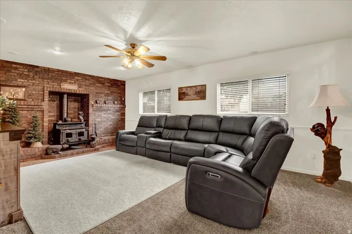 Spacious living room featuring a ceiling fan with light fixture, exposed brick fireplace with built-in hearth, expansive windows with plantation shutters, neutral carpeting, and recessed lighting throughout