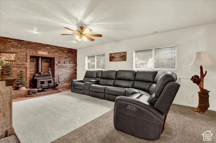 Spacious living room featuring a ceiling fan with light fixture, exposed brick fireplace with built-in hearth, expansive windows with plantation shutters, neutral carpeting, and recessed lighting throughout