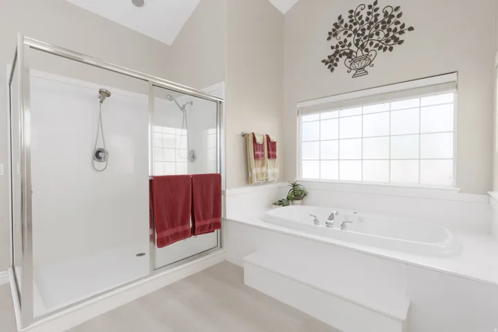 Vaulted ceiling with an integrated recessed light, light-toned flooring, glass-enclosed shower with a handheld showerhead, built-in soaking tub, and a large window with white grilles