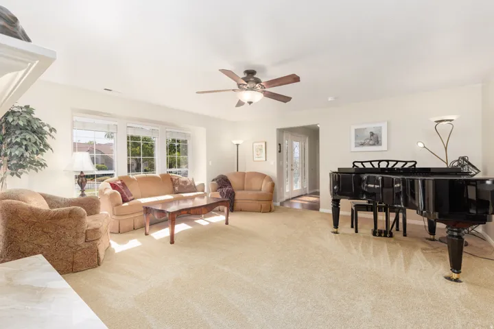 Spacious living room featuring gleaming wood-finish flooring, abundant natural light from multiple French doors and windows, a ceiling fan with integrated lighting, and a grand piano