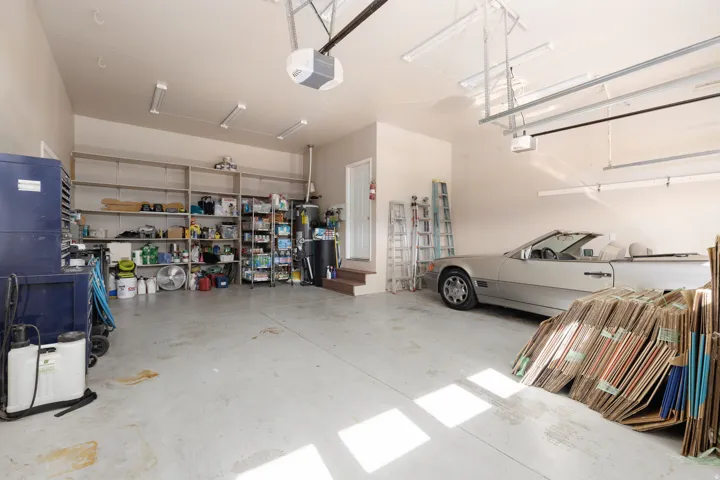 Spacious garage with epoxy-finished concrete flooring, abundant natural light from multiple skylights and garage doors, extensive wall-mounted shelving and storage systems, and room for multiple vehicles alongside workshop and material storage