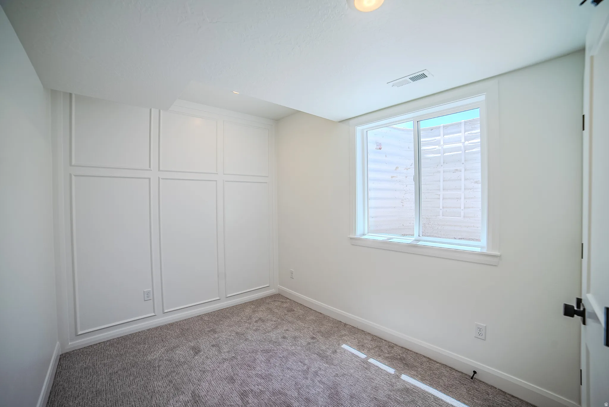 White paneled wall detailing, light gray carpet flooring, egress window with white trim, recessed lighting, and a white interior door with black hardware