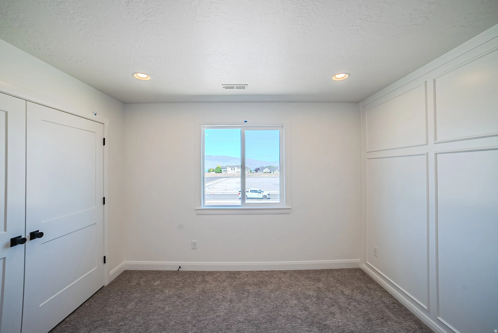 White interior walls, recessed ceiling lighting, a window with exterior scenery, paneled bifold doors with black hardware, and an accent wall featuring wainscoting