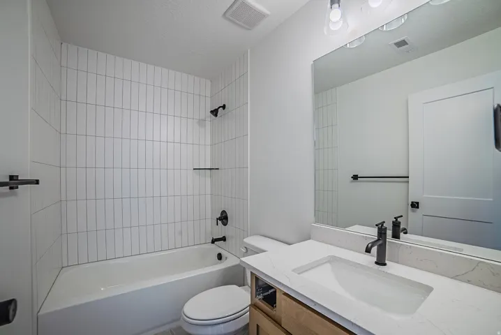 Bathroom featuring a tiled shower surround, built-in bathtub, wood-finish vanity with an integrated sink, quartz countertop, and black hardware