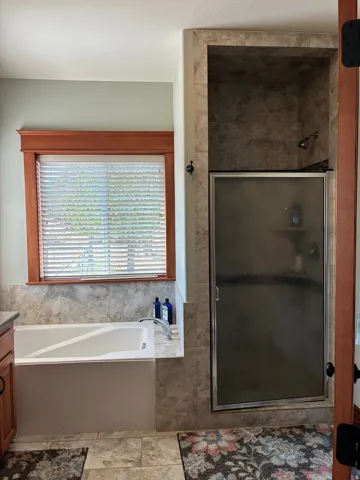 Primary Bathroom featuring a soaking tub with tile surround, large window with wood-finish frame and privacy blinds, ceramic tile flooring, and an enclosed shower with glass enclosure and tile walls
