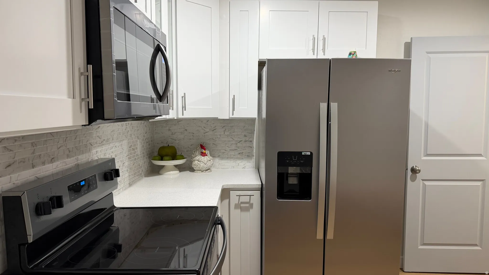 Modern kitchen featuring stainless steel side-by-side refrigerator with ice and water dispenser, black electric range and microwave, white cabinetry with brushed nickel hardware, and gray subway tile backsplash above white quartz countertops