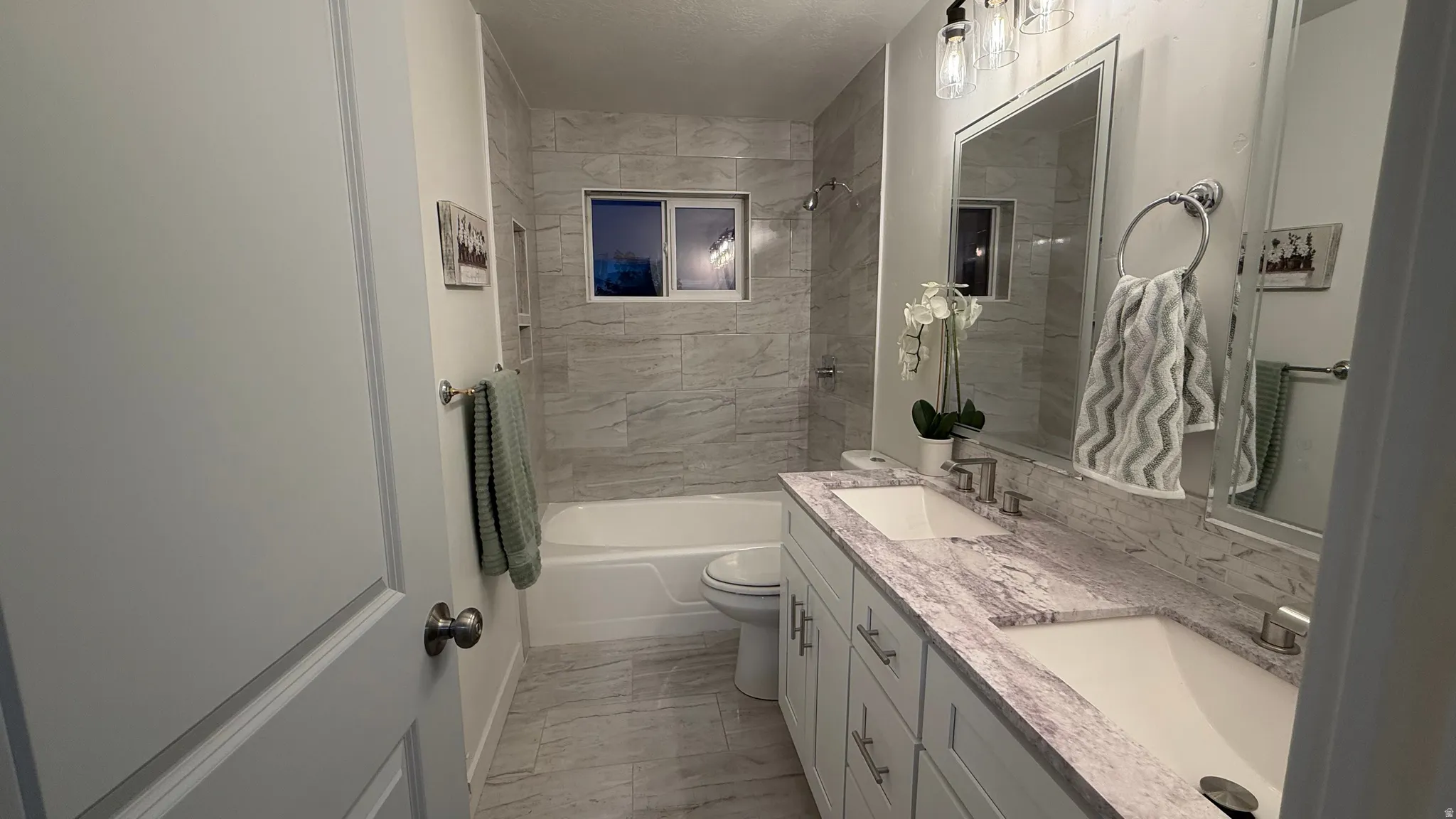 Modern bathroom featuring dual vanity with white cabinetry and marble-look countertops, large framed mirror with integrated lighting, walk-in shower with tile surround and glass enclosure, and stone-finish flooring throughout
