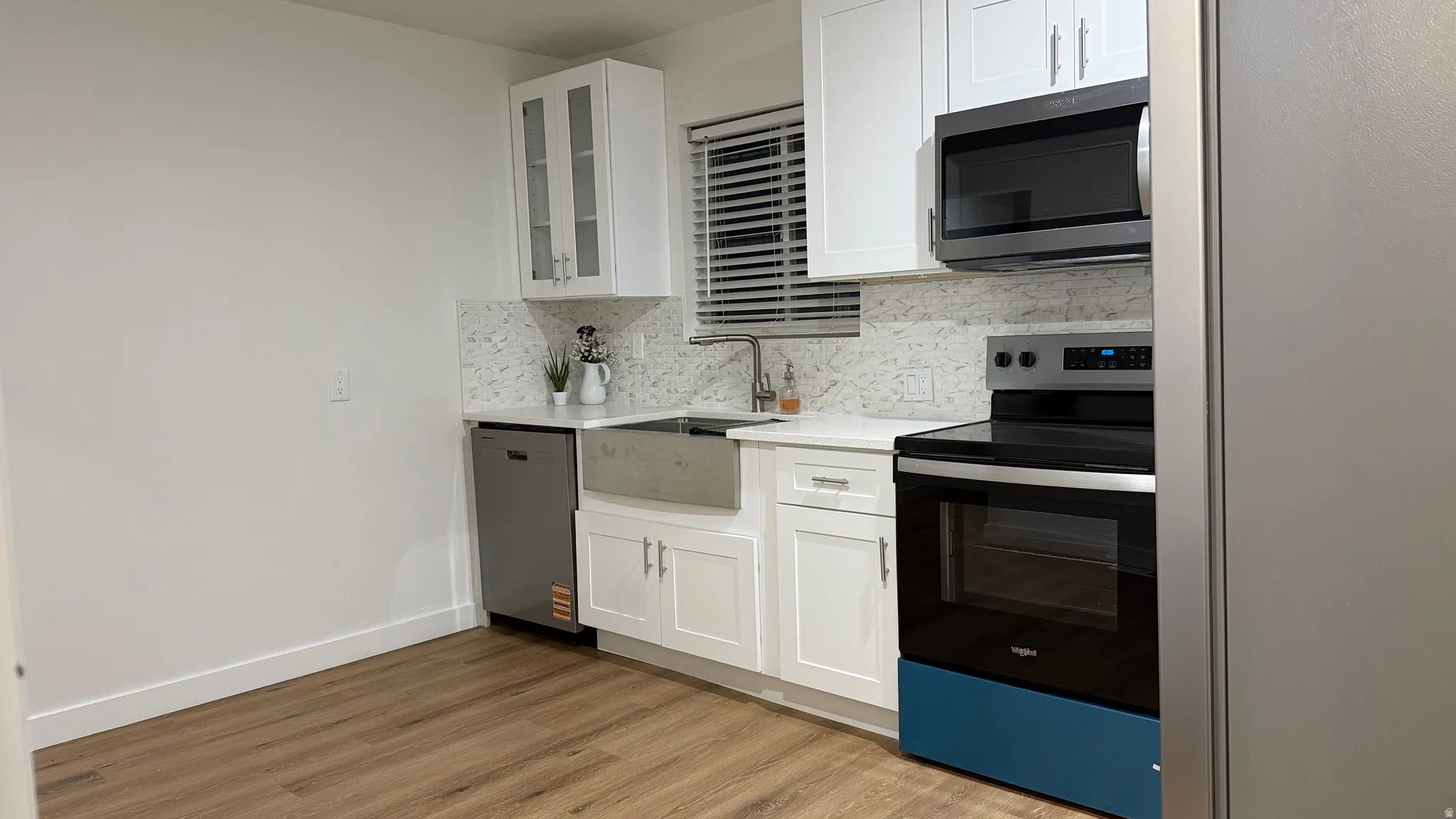 Wood-finish flooring, white cabinetry with display glass, stainless steel farmhouse sink, white countertops, and a full tile backsplash