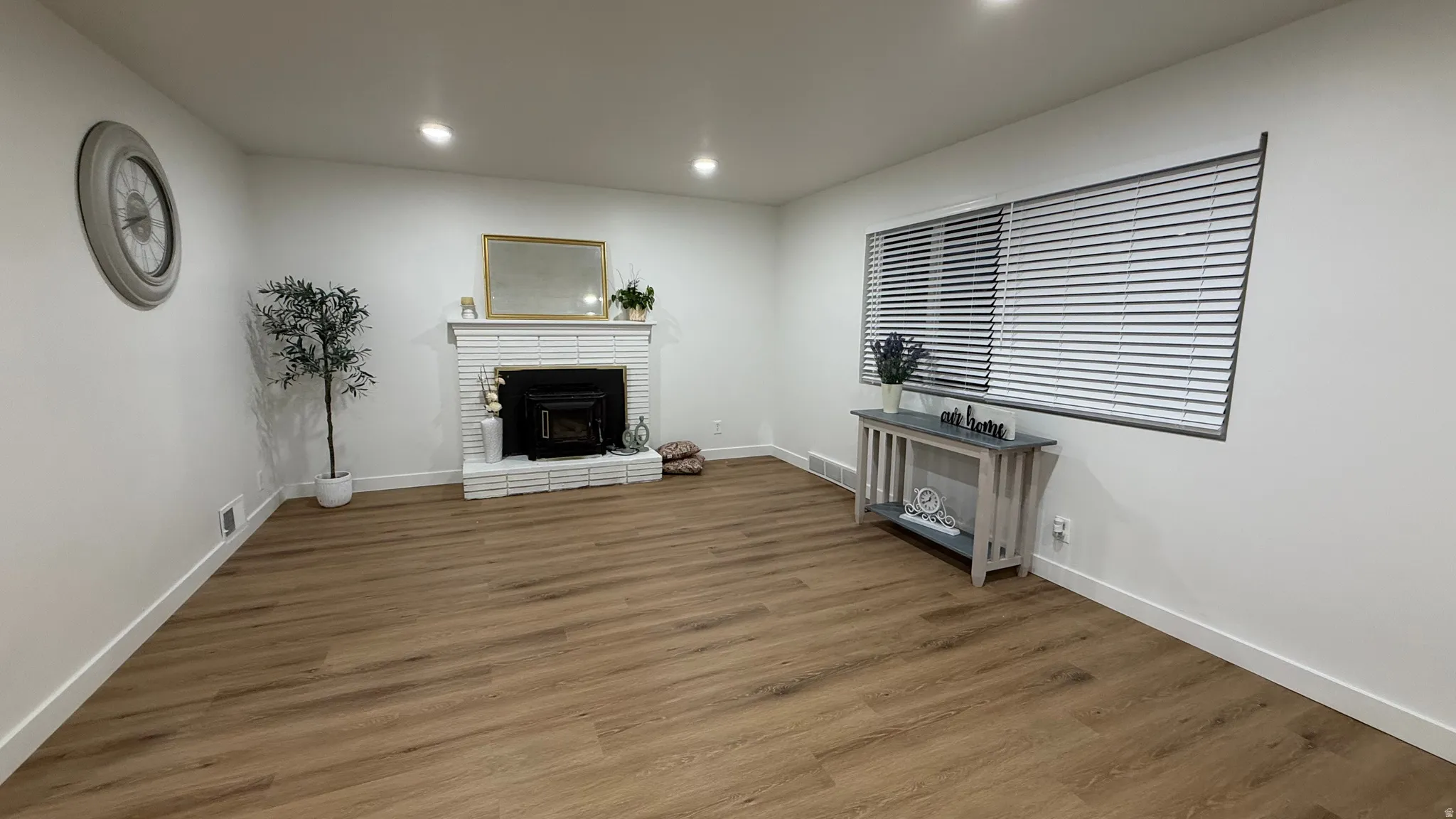 White brick fireplace with mantel, wood-finish flooring, recessed lighting, and horizontal blinds