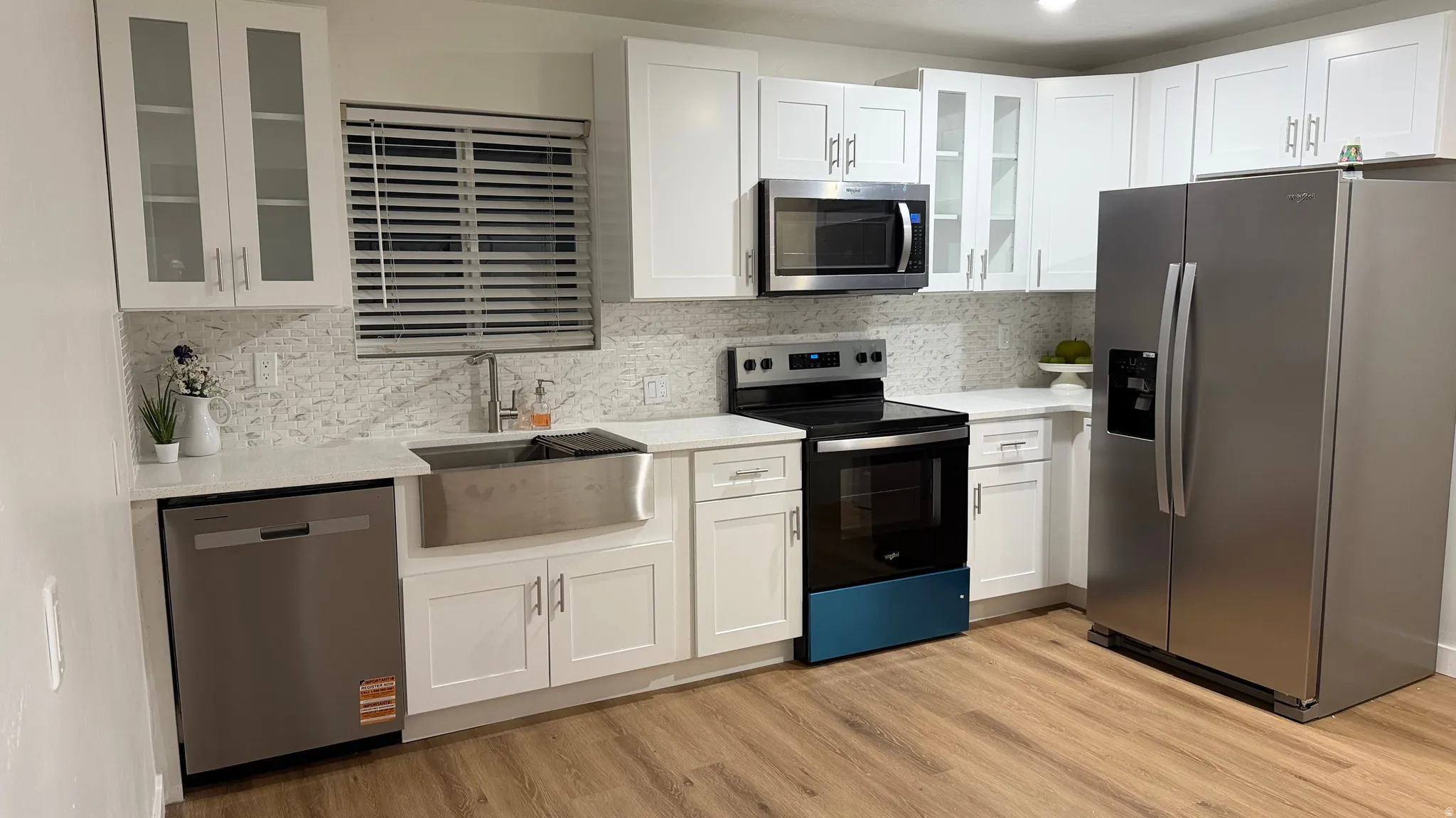 Modern kitchen featuring white cabinetry with glass-front upper cabinets, stainless steel appliances including a French-door refrigerator and electric range, a farmhouse-style apron sink with brushed nickel fixtures, quartz countertops with a neutral mosa