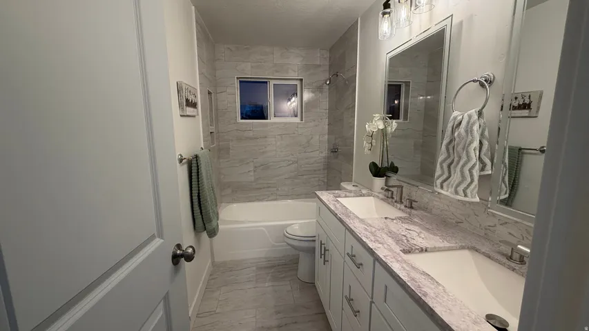 Modern bathroom featuring dual vanity with white cabinetry and marble-look countertops, large framed mirror with integrated lighting, walk-in shower with tile surround and glass enclosure, and stone-finish flooring throughout
