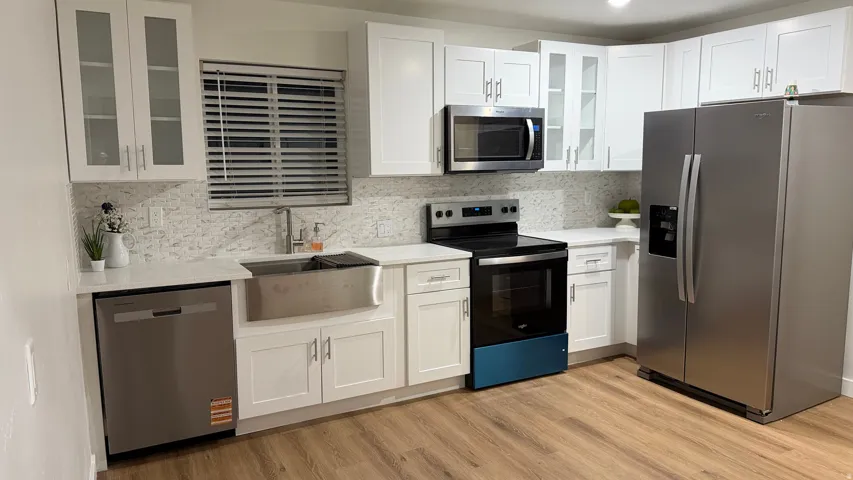 Modern kitchen featuring white cabinetry with glass-front upper cabinets, stainless steel appliances including a French-door refrigerator and electric range, a farmhouse-style apron sink with brushed nickel fixtures, quartz countertops with a neutral mosa