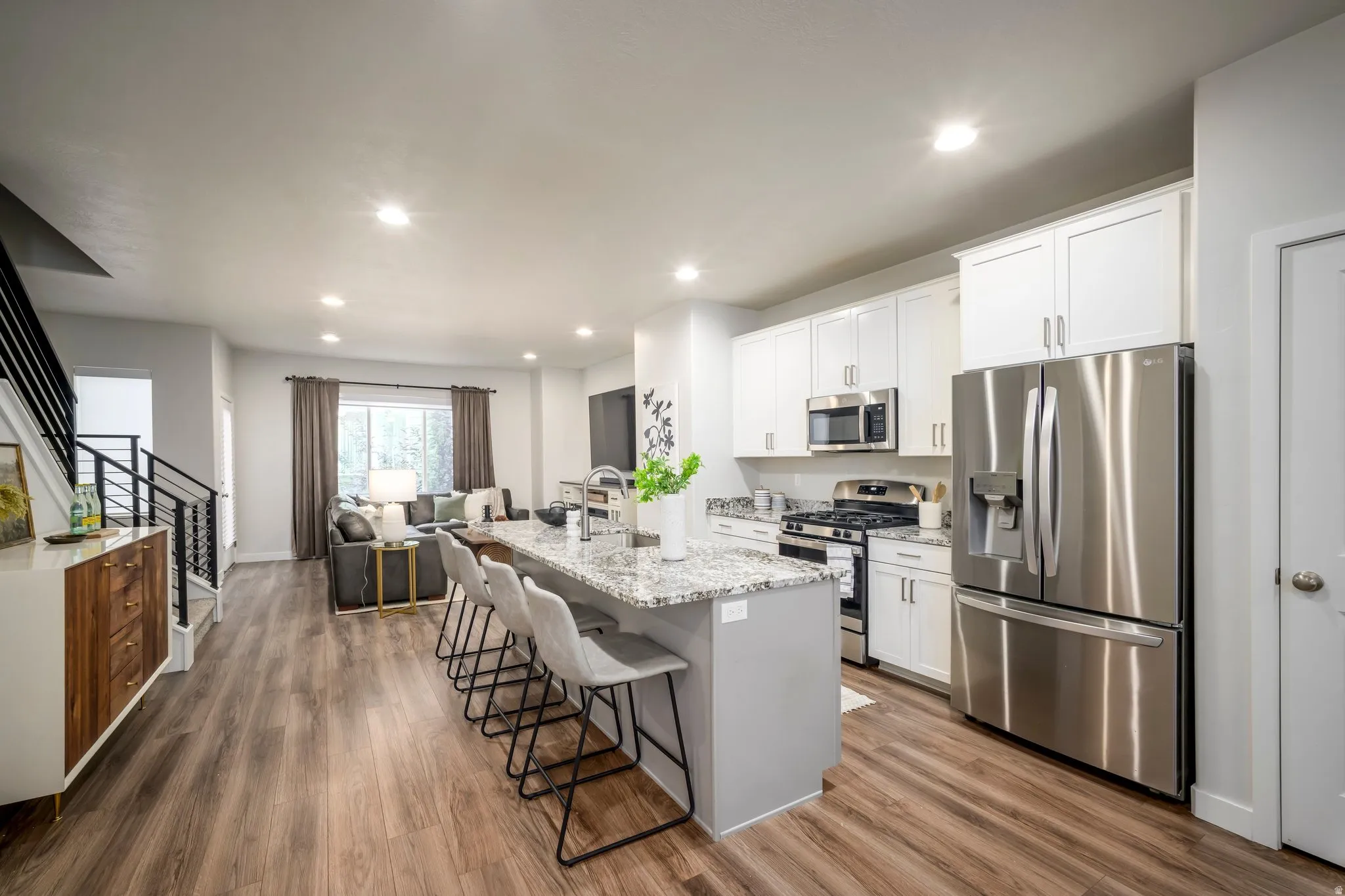 Open-concept living area featuring wood-finish flooring, recessed lighting, and a kitchen with white cabinetry, stainless steel appliances, and a generous, granite island