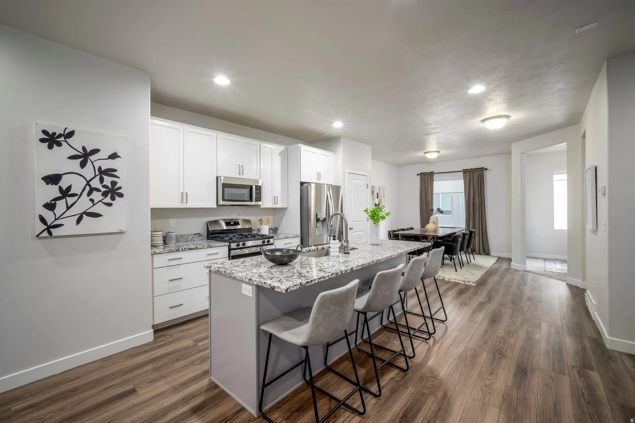 Modern kitchen and dining area featuring white cabinetry with stainless steel appliances, granite-style countertops, a center island with seating for multiple guests, recessed and pendant lighting, and wood-finish flooring throughout the open-concept spac
