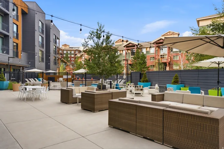 Expansive outdoor entertaining space featuring multiple seating areas with woven lounge furniture, concrete paving throughout, shade umbrellas, built-in planters with mature landscaping, and modern residential architecture surrounding the courtyard