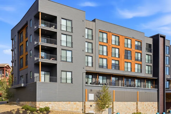 Multi-story building featuring gray siding, wood-finish paneling, multiple balconies with glass railings, and a stone-finish base