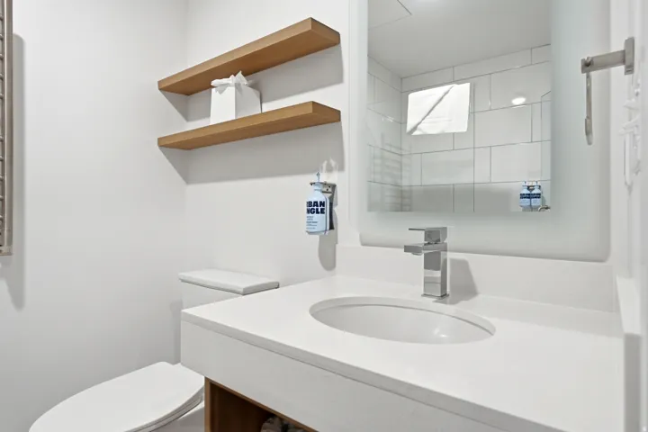 Integrated single basin sink with chrome faucet, floating wood-finish shelving, wall-mounted mirror, and a white toilet