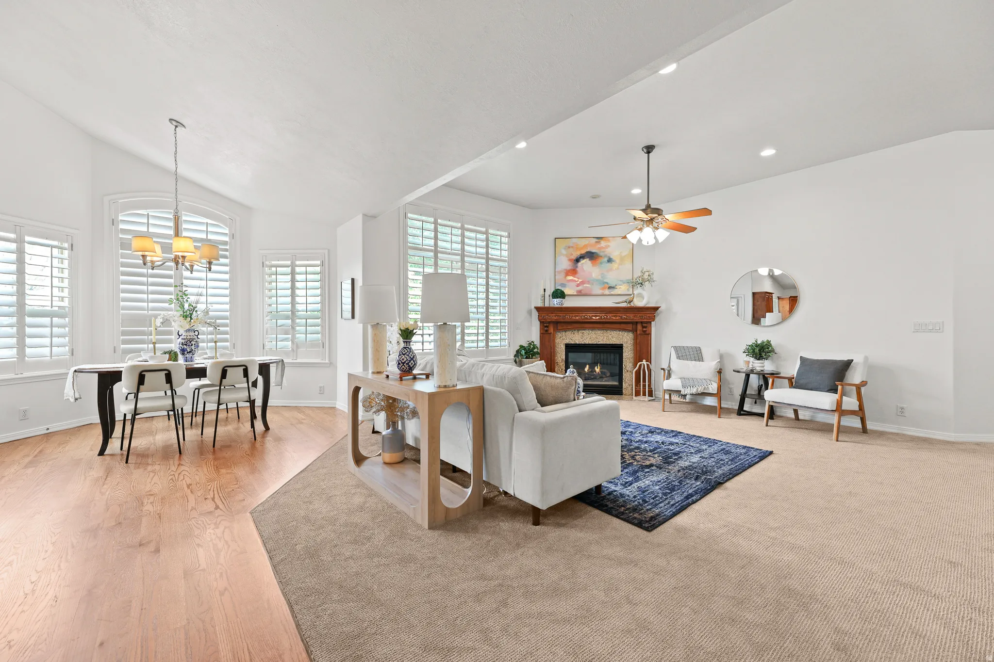 Vaulted ceilings, wood-finish flooring, plantation shutters, gas fireplace with a decorative mantel, and a ceiling fan with integrated lighting
