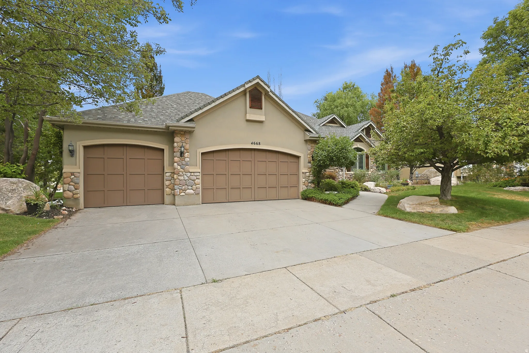 Stucco exterior with stone accents, two arched garage doors, concrete driveway, and mature landscaping