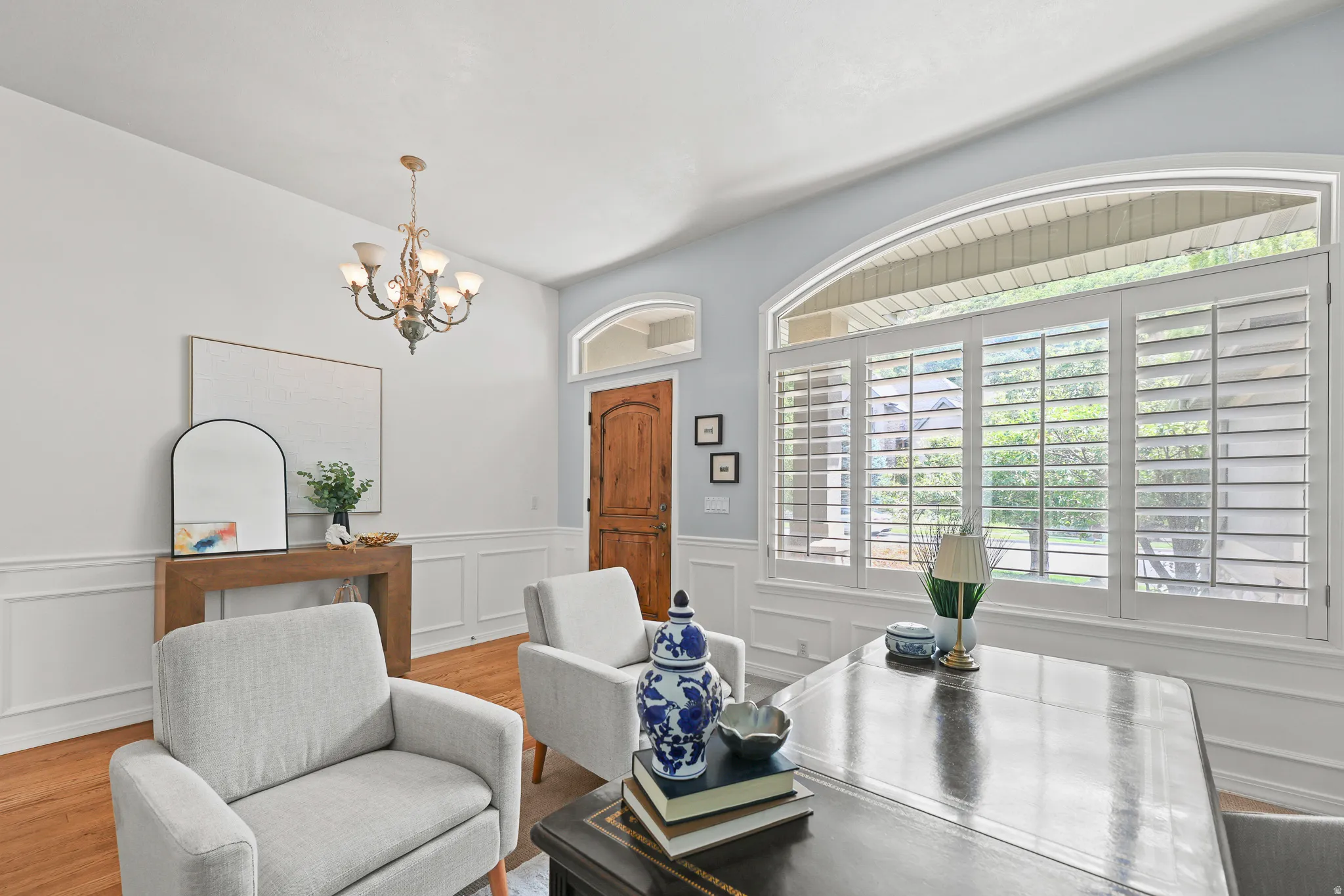 Arched window with plantation shutters, wood-finish flooring, wainscoting, wood panel door, and an ornate chandelier