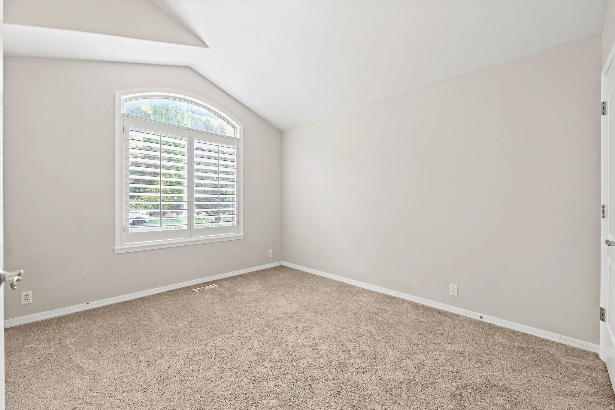 Arched window with plantation shutters, vaulted ceiling, neutral wall color, and wall-to-wall carpeting