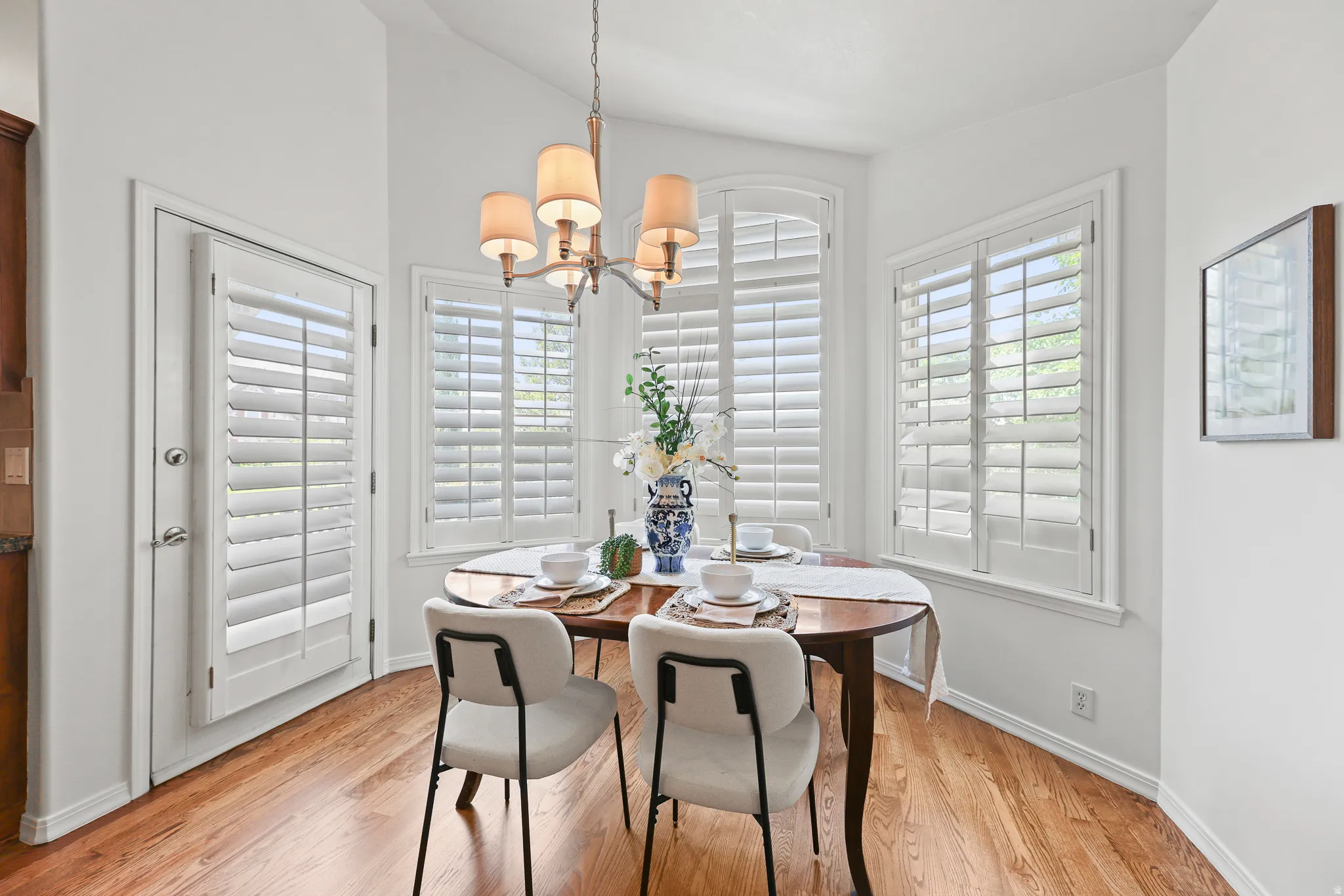Hardwood flooring with a semicircular dining area, multiple windows with white plantation shutters, and a chandelier with fabric shades