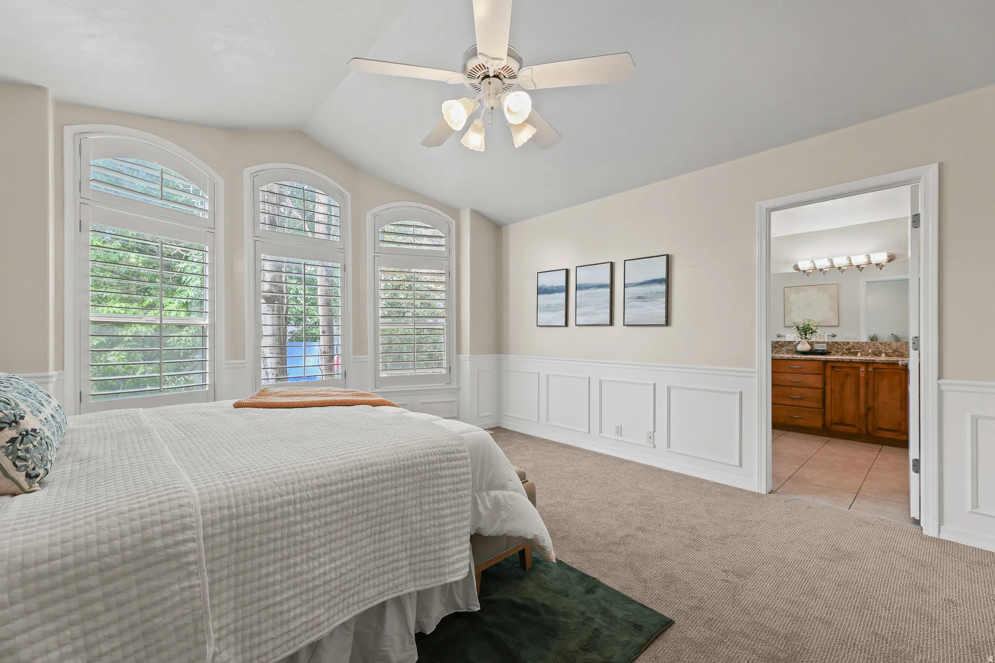 Vaulted ceiling with a ceiling fan, arched windows featuring plantation shutters, wainscoting, and carpet flooring