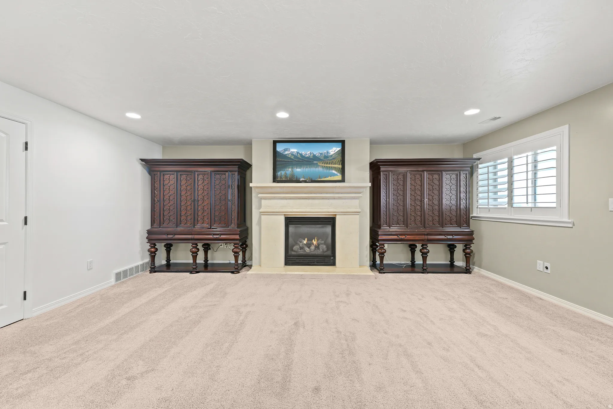 Gas fireplace with a stone surround and mantel, recessed lighting, wall-to-wall carpeting, and a window with plantation shutters