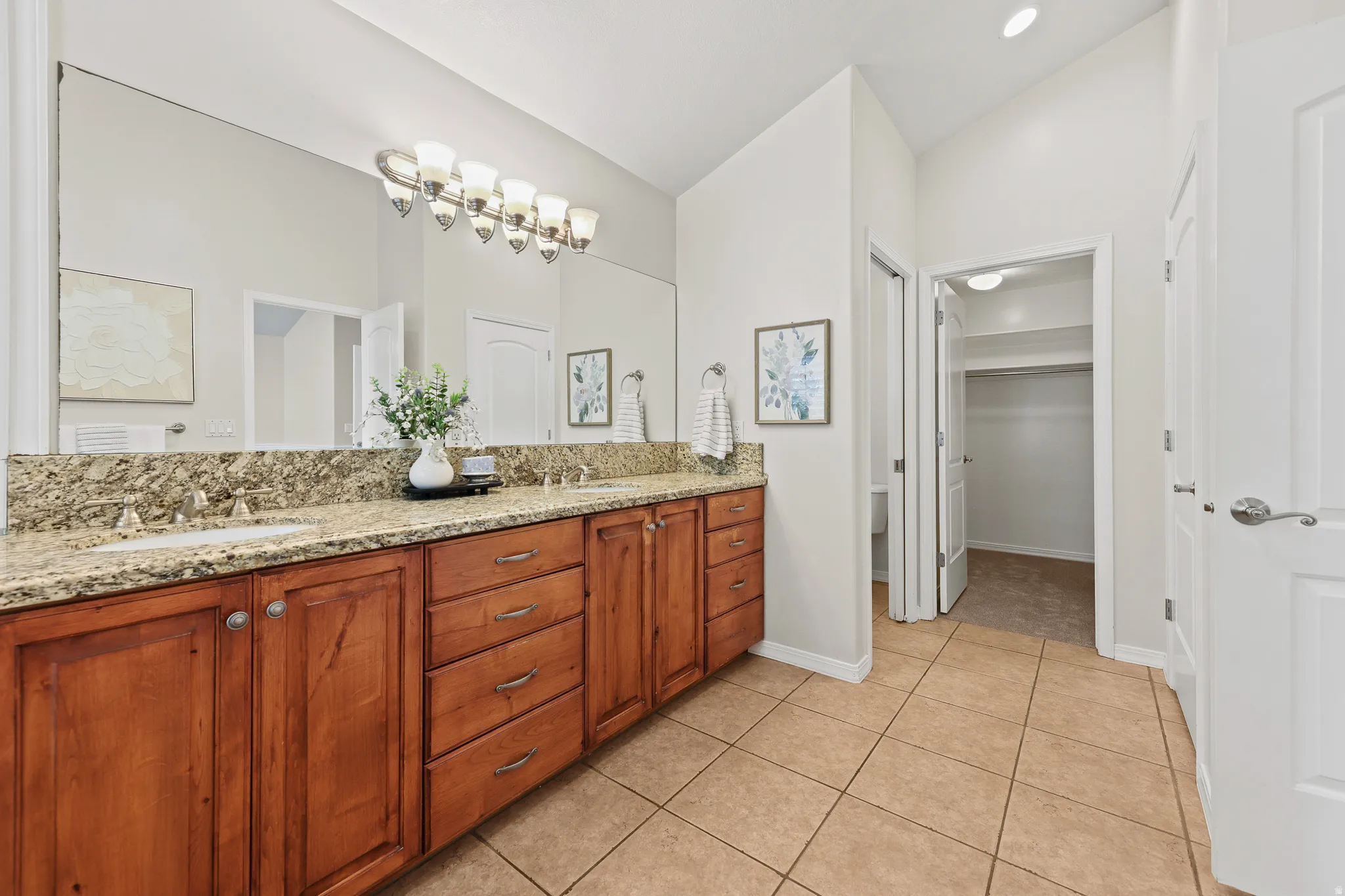 Dual sinks with granite countertop, wood-finish cabinetry, tile flooring, and vaulted ceiling