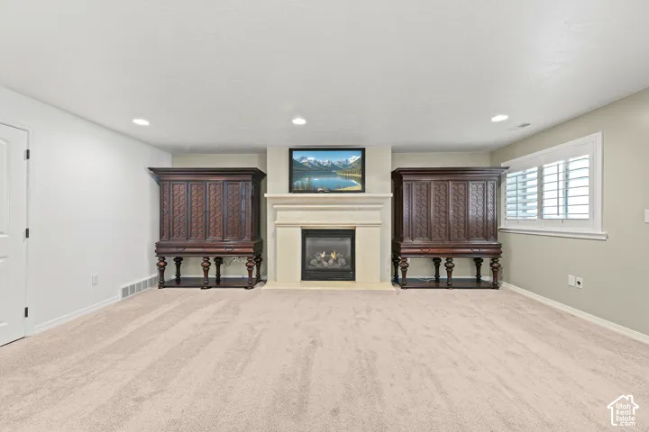 Gas fireplace with a stone surround and mantel, recessed lighting, wall-to-wall carpeting, and a window with plantation shutters
