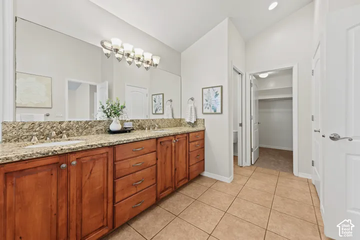 Dual sinks with granite countertop, wood-finish cabinetry, tile flooring, and vaulted ceiling
