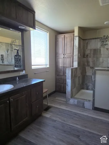 Wood-finish flooring, dark wood cabinetry, built-in storage cabinet, tiled walk-in shower, and a single vanity with a solid surface countertop