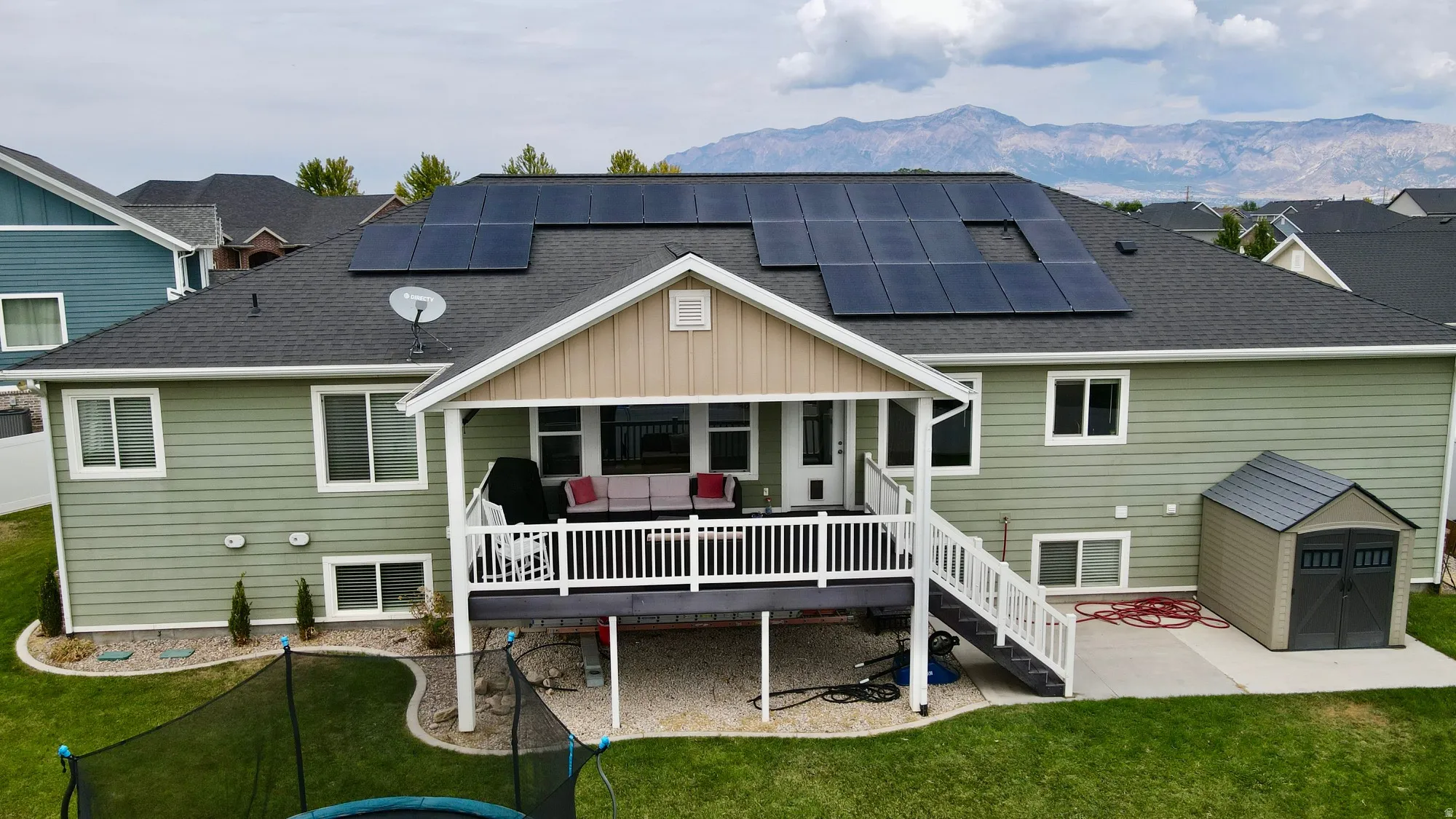 Spacious home featuring a covered deck with white railings, solar panel array on the roof, composite or asphalt shingle roofing, multi-pane windows throughout, and manicured lawn with mature landscaping and mountain views in the distance