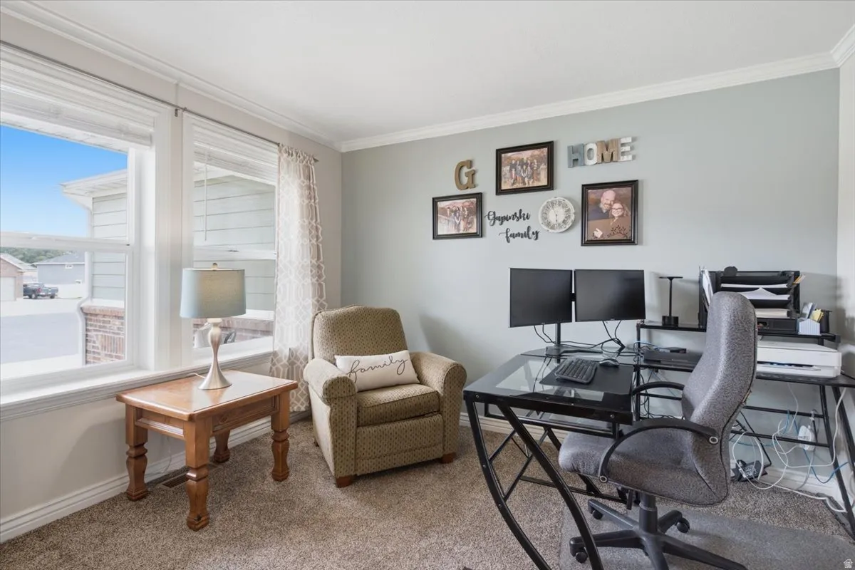 Bright, multi-purpose room featuring large windows with natural light, neutral wall finishes, built-in desk workspace with dual monitor setup, and carpeted flooring throughout