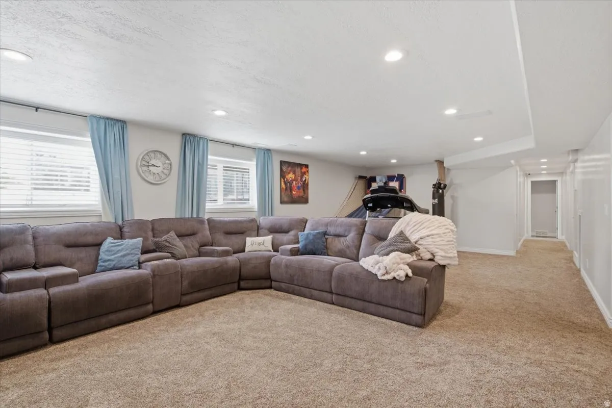 Spacious recreational room featuring a sectional layout with ample seating, recessed ceiling lighting, neutral carpet flooring, and large windows with blue drapery allowing natural light throughout the space