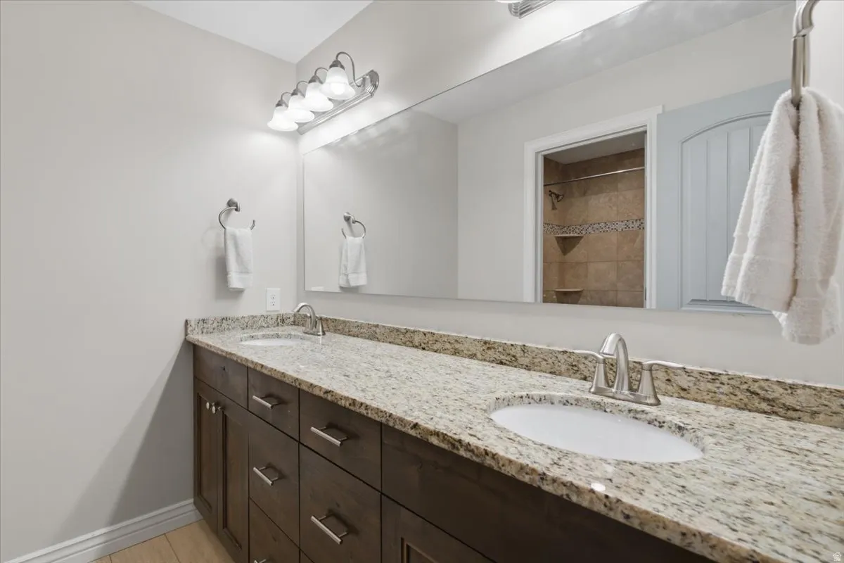 Spa-inspired double vanity featuring a granite-look countertop with integrated sinks, dark wood cabinetry with chrome hardware, and a large frameless mirror with overhead and task lighting