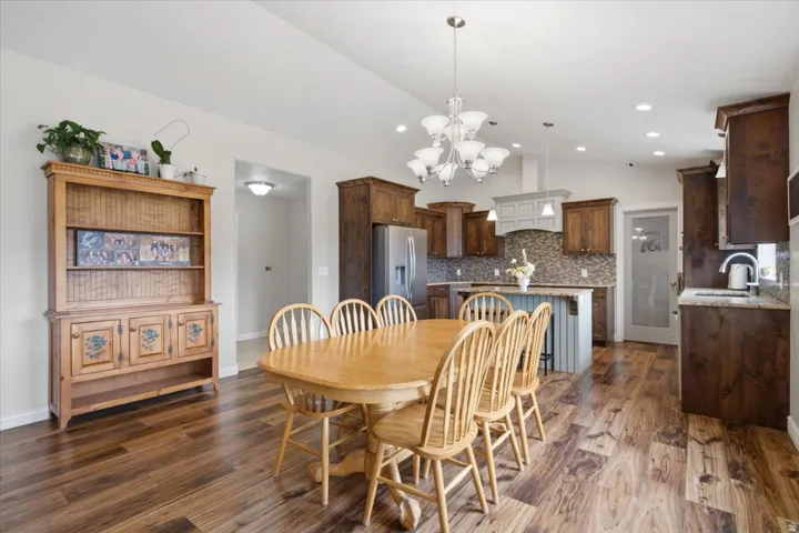 Open-concept dining and kitchen area featuring hardwood flooring, a wood dining table with seating for six, custom cabinetry with warm wood finishes, stainless steel appliances, stone countertops, a multi-pendant light fixture, and recessed lighting throu