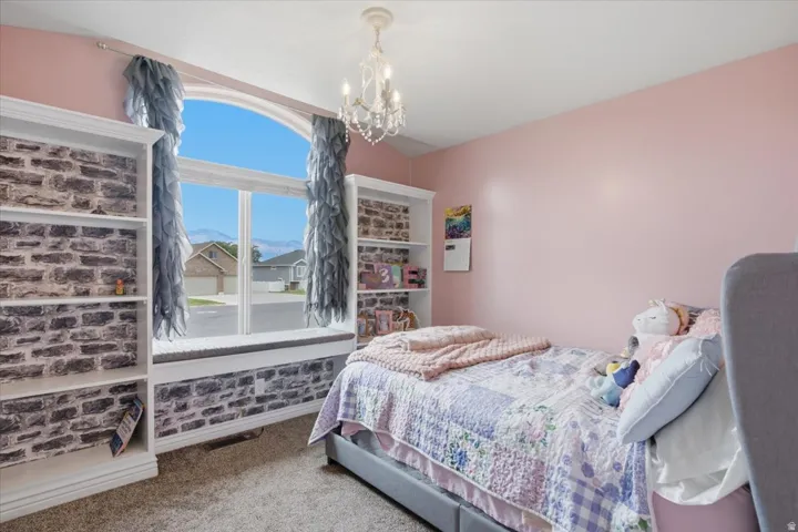 Bright, light-filled bedroom featuring a large window with brick surround and decorative hardware, soft pink walls, carpeted flooring, a crystal chandelier, and built-in shelving with display space