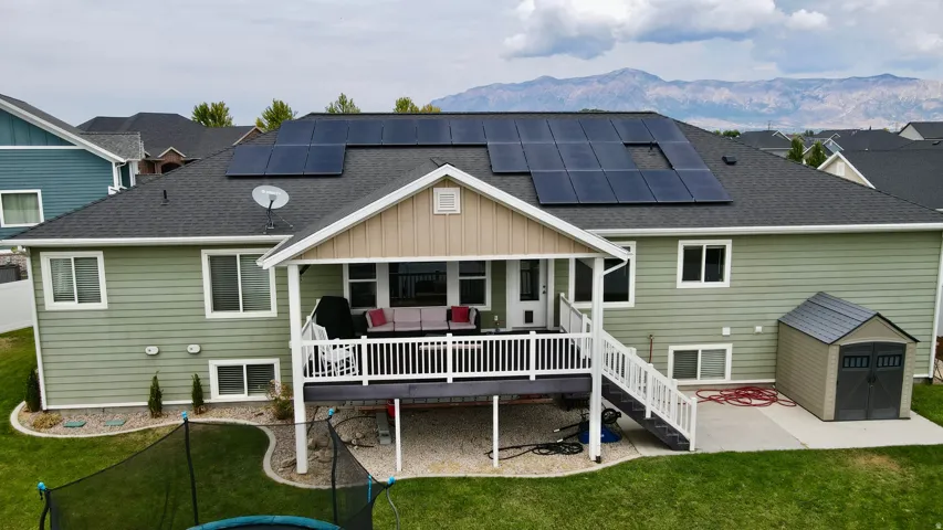 Spacious home featuring a covered deck with white railings, solar panel array on the roof, composite or asphalt shingle roofing, multi-pane windows throughout, and manicured lawn with mature landscaping and mountain views in the distance