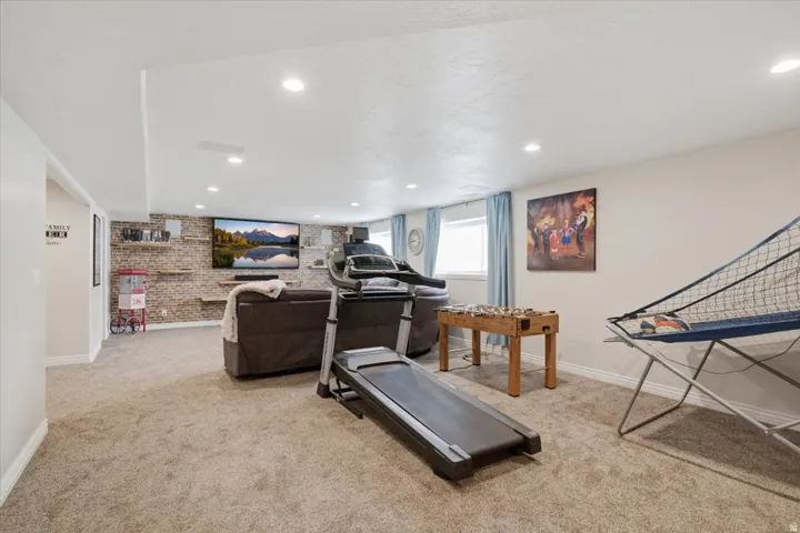Finished basement recreation room featuring neutral carpet flooring, recessed ceiling lighting, brick accent wall with built-in shelving, and large windows providing natural light