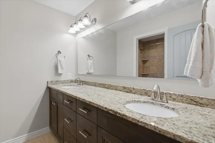 Spa-inspired double vanity featuring a granite-look countertop with integrated sinks, dark wood cabinetry with chrome hardware, and a large frameless mirror with overhead and task lighting