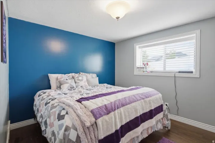 Bedroom featuring a striking blue accent wall, wood-finish flooring, and a large window with horizontal blinds providing natural light and privacy