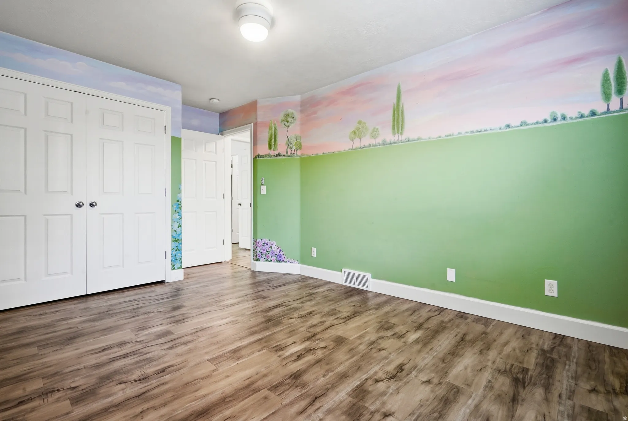 Spacious room featuring wood-finish flooring, fresh green accent walls with scenic mural detail, white six-panel closet doors, and multiple electrical outlets