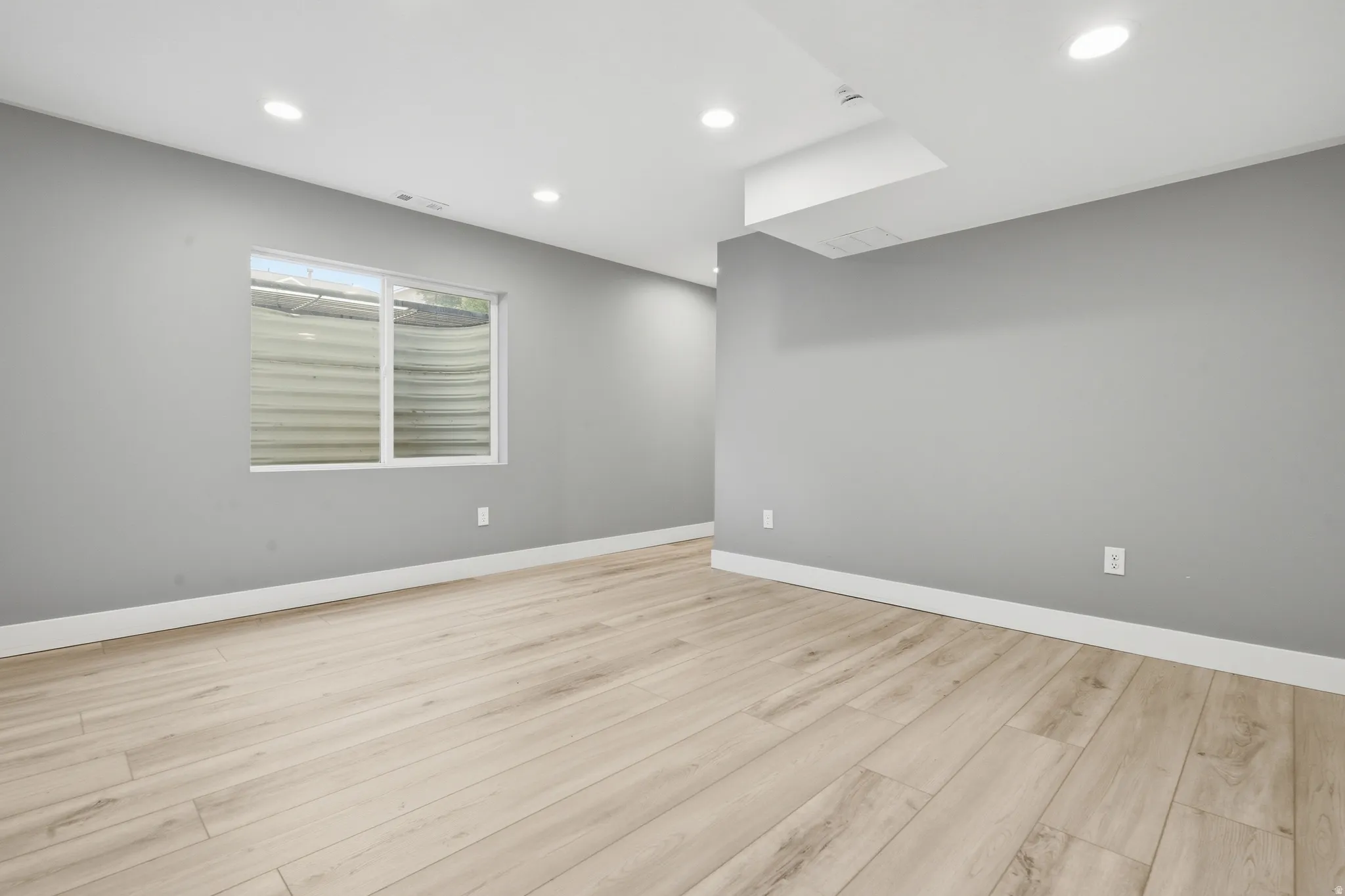 Spacious room featuring light wood-finish flooring, gray painted walls, recessed ceiling lighting, and a skylight providing natural illumination